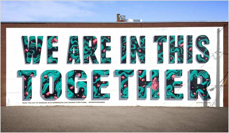 White mural on a brick wall with the words We Are in This Together, in large all caps letters. The letters are filled with a turquoise and black swirl pattern with pink cubes and other shapes. The subtitle reads: 'What to say to someone with depression can change everything. # find your words. lets talk co . org