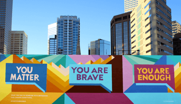 Freestanding mural in front of a cityscape. The mural features three rectangular speech bubbles with all caps text You Matter, You Are Brave and You Are Enough in them, over a geometric background of intersecting concentric triangles in shades of orange, green, blue and burgundy.